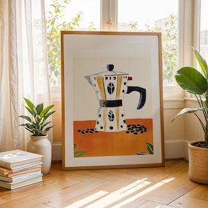 May include: A framed art print of a coffee maker in a kitchen setting. The artwork features a yellow, black, and white coffee maker design with coffee beans on an orange background. The print is displayed on a wooden floor near plants and books.
