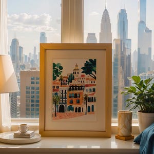 May include: A framed print of a colourful illustration depicting a cityscape with buildings, palm trees, and a blue sky. The print is in a light wood frame and is sitting on a white surface.