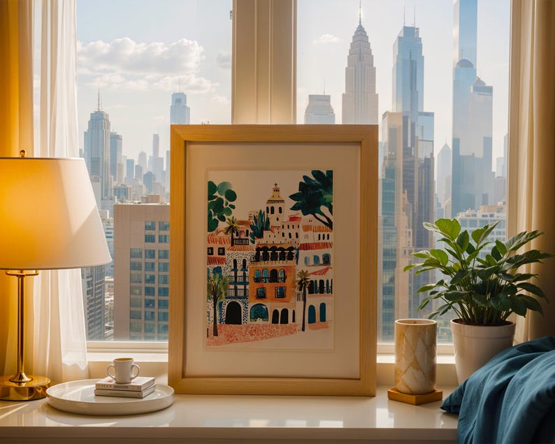 May include: A framed print of a colourful illustration depicting a cityscape with buildings, palm trees, and a blue sky. The print is in a light wood frame and is sitting on a white surface.