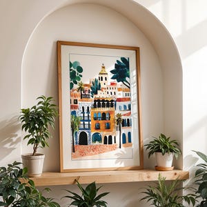 May include: A framed watercolour painting of a colourful cityscape with buildings, trees, and a church steeple. The painting is in a light wood frame and is hanging on a wall.
