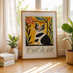 May include: Framed art print featuring a stylized cat illustration in black, white, and orange against a yellow background. The print includes the phrase "C'est la vie" in a classic font. The artwork is displayed in a room with plants and natural light.