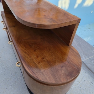 May include: A mid-century modern style wooden cabinet with a curved design. The cabinet is made of dark brown wood with a glossy finish. It features gold-colored drawer pulls and a black base. The cabinet has a unique, rounded shape.