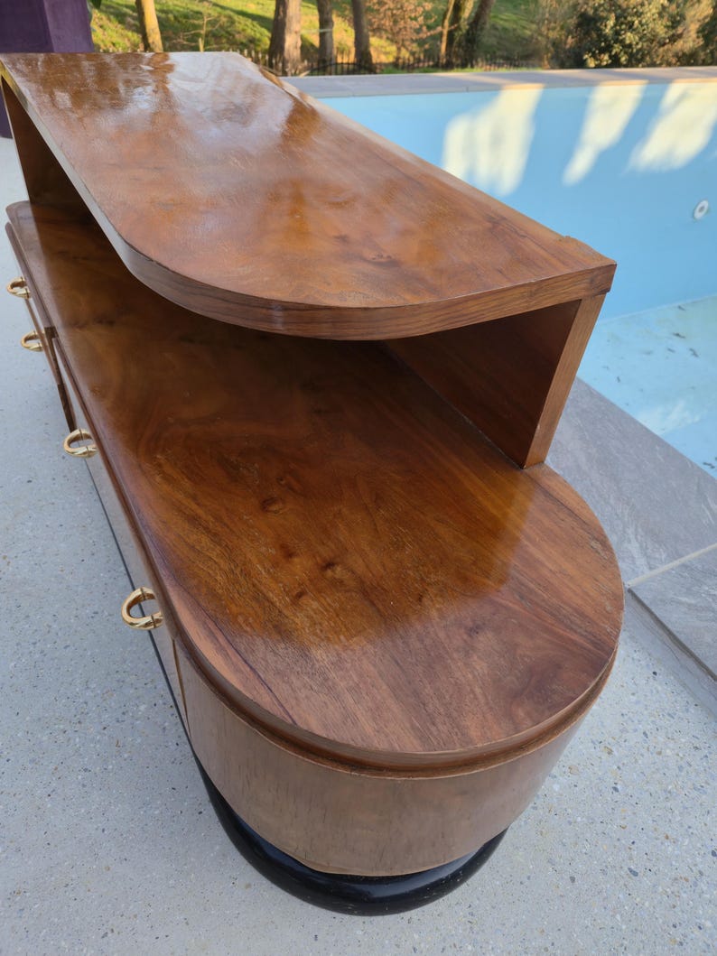 May include: A mid-century modern style wooden cabinet with a curved design. The cabinet is made of dark brown wood with a glossy finish. It features gold-colored drawer pulls and a black base. The cabinet has a unique, rounded shape.