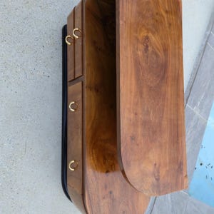 May include: A wooden cabinet with a curved design and a rich, brown finish. The cabinet features three small drawers with gold-colored, circular handles and a large, open compartment. The cabinet has a black trim on the side.