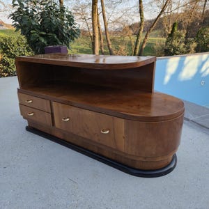 May include: A mid-century modern wooden cabinet with a curved design. The cabinet has a dark wood finish, two drawers, and gold-colored handles. It sits on a black base and has a shelf on top.