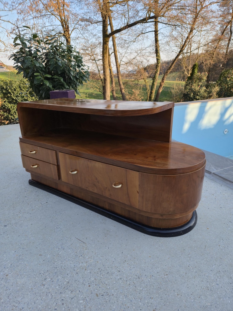 May include: A mid-century modern wooden cabinet with a curved design. The cabinet has a dark wood finish, two drawers, and gold-colored handles. It sits on a black base and has a shelf on top.