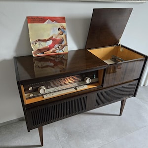May include: A retro wooden console stereo unit with a record player and radio. The dark brown cabinet features a hinged lid, a speaker grille, and a control panel with knobs. An album cover with a woman in a red swimsuit is on top.