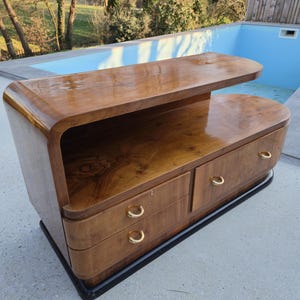 May include: A mid-century modern style wooden cabinet with a curved design. The cabinet has two drawers with gold-colored handles and a top shelf. The wood has a rich, warm tone and a glossy finish. The base is black.