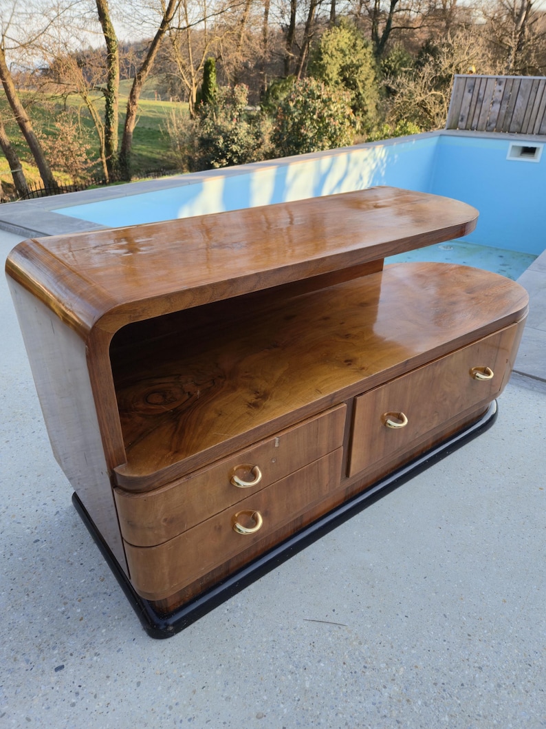 May include: A mid-century modern style wooden cabinet with a curved design. The cabinet has two drawers with gold-colored handles and a top shelf. The wood has a rich, warm tone and a glossy finish. The base is black.
