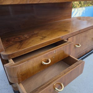 May include: A vintage wooden dresser with a glossy finish. The dresser has two open drawers with gold-colored handles. The top of the dresser has a shelf. The wood grain is visible. The dresser is a warm brown color.