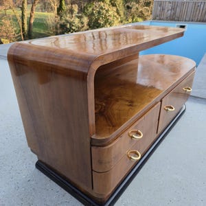 May include: A mid-century modern style wooden nightstand with a curved design. The nightstand has a dark wood finish, three drawers with gold-colored handles, and a black base. The top surface is smooth and reflective.