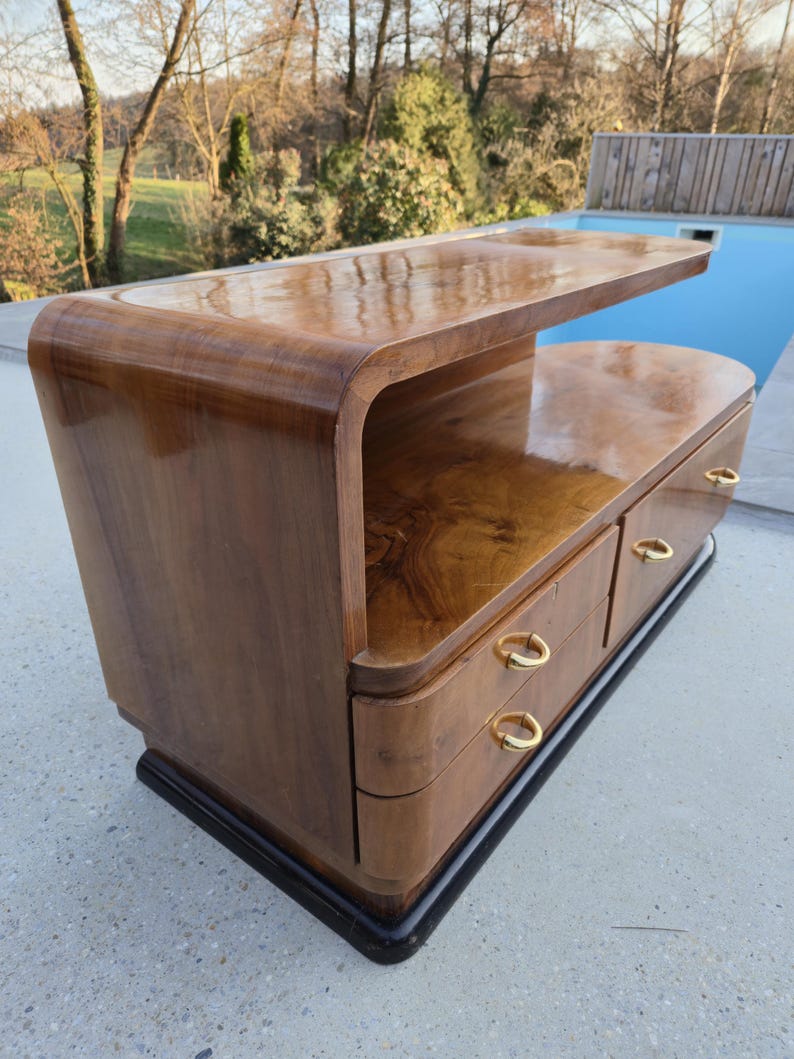 May include: A mid-century modern style wooden nightstand with a curved design. The nightstand has a dark wood finish, three drawers with gold-colored handles, and a black base. The top surface is smooth and reflective.
