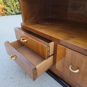 May include: A vintage wooden cabinet with two open drawers and a top shelf. The cabinet is made of a warm-toned wood with a glossy finish. The drawers have gold-colored, curved handles. The cabinet sits on a black base.