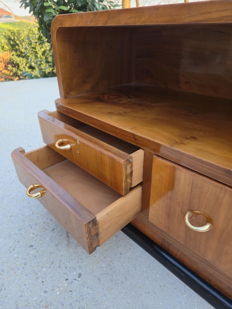 May include: A vintage wooden cabinet with two open drawers and a top shelf. The cabinet is made of a warm-toned wood with a glossy finish. The drawers have gold-colored, curved handles. The cabinet sits on a black base.