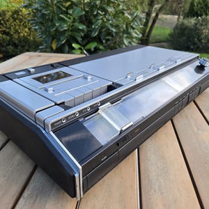 Op de afbeelding: Een vintage zilver-zwarte stereo cassette speler en recorder. Het apparaat heeft een strak, rechthoekig ontwerp met meerdere knoppen, een cassettesleuf en een doorzichtige plastic hoes. Het apparaat staat op een houten oppervlak.