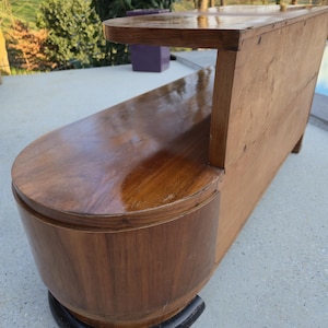 May include: A vintage wooden bench with a curved seat and backrest. The bench is made of dark brown wood with a glossy finish. The base is a rounded shape with a black trim. The bench has a small shelf on the back.