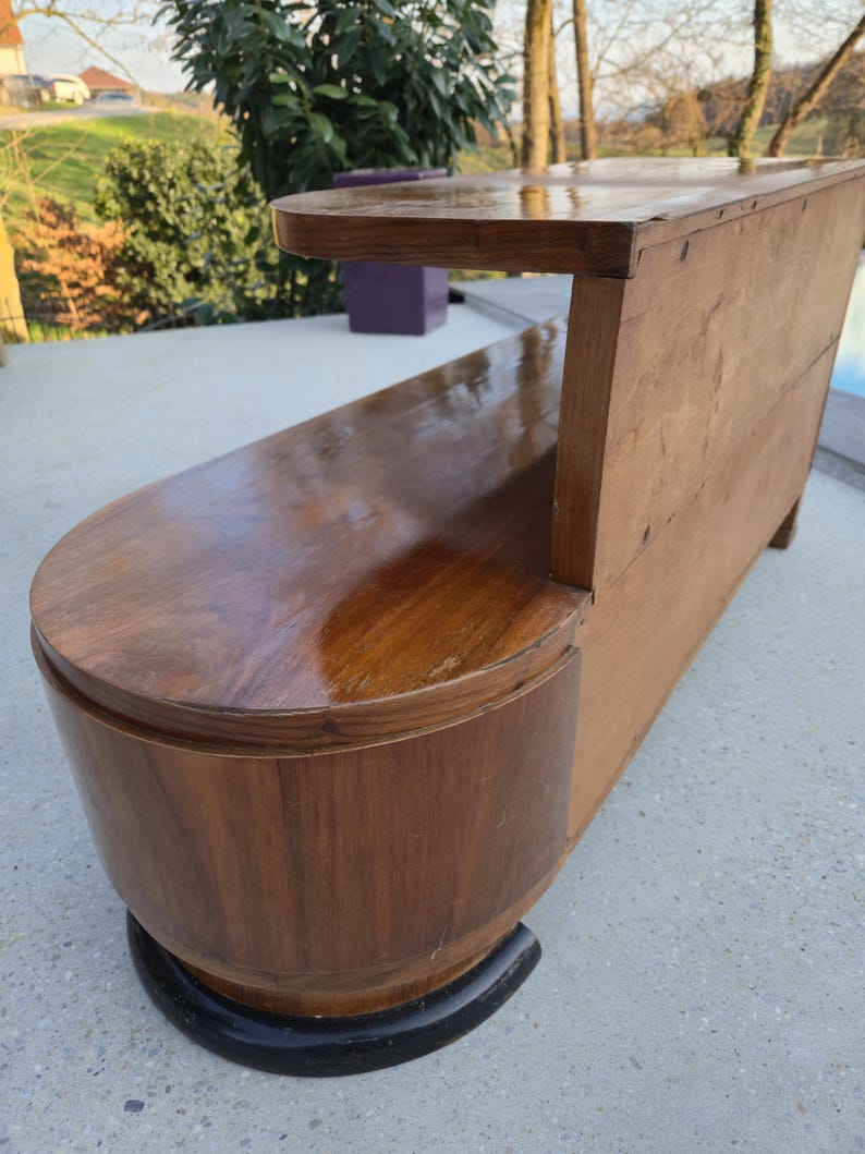 May include: A vintage wooden bench with a curved seat and backrest. The bench is made of dark brown wood with a glossy finish. The base is a rounded shape with a black trim. The bench has a small shelf on the back.
