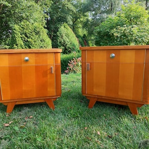 May include: Two wooden bedside tables with a single drawer and a door. The bedside tables have a light wood finish and are sitting on a green lawn.