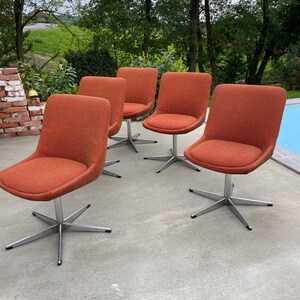 Peut inclure: Quatre chaises pivotantes avec un rev&ecirc;tement en tissu orange et des bases chrom&eacute;es. Les chaises sont dispos&eacute;es en rang&eacute;e sur une terrasse en b&eacute;ton.