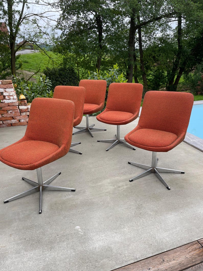 Peut inclure: Quatre chaises pivotantes avec un rev&ecirc;tement en tissu orange et des bases chrom&eacute;es. Les chaises sont dispos&eacute;es en rang&eacute;e sur une terrasse en b&eacute;ton.