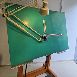 May include: An antique drafting table with a green drawing surface and an adjustable light fixture. The table has an orange wooden base and a black footrest. The drawing surface has a mechanical arm for precise drawing.