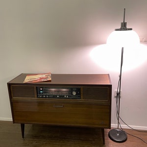 May include: A vintage wooden record player console with a record on top, a drawer, and a radio tuner. The console is in a room with a white floor lamp.