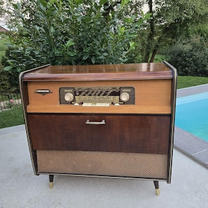 Könnte beinhalten: Vintage Holz-Radiokonsole mit hellem Holzdeckel und dunklem Holzschrank. Das Radio verfügt über einen Lautsprecher, Drehregler und Tasten. Die Konsole hat goldfarbene Beine und eine gewebte Lautsprecherabdeckung.