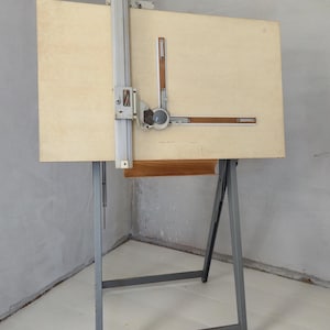 May include: A drafting table featuring a large, rectangular, light-colored wooden board mounted on a gray metal frame. The board includes an adjustable vertical and horizontal drafting machine mechanism with a circular central dial and metal rails.