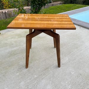 May include: A small, square wooden table with a light brown top and four angled legs. The table is made of wood with a natural grain pattern.
