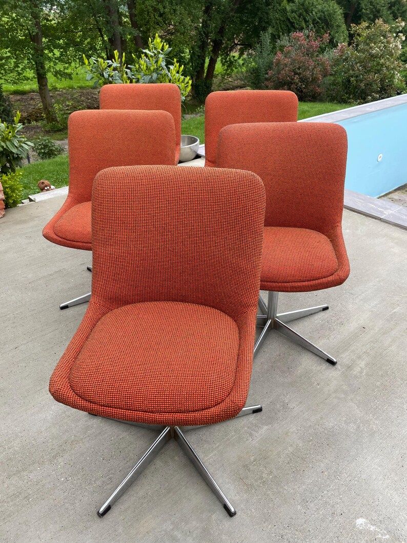 Peut inclure: Quatre chaises pivotantes orange avec des bases chrom&eacute;es. Les chaises sont dispos&eacute;es en demi-cercle sur une terrasse en b&eacute;ton.