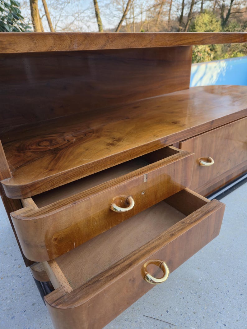 May include: A vintage wooden dresser with a glossy finish. The dresser has two open drawers with gold-colored handles. The top of the dresser has a shelf. The wood grain is visible. The dresser is a warm brown color.