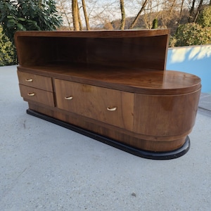 May include: A mid-century modern wooden cabinet with a curved design. The cabinet has a dark wood finish, three drawers with gold-colored handles, and a recessed top shelf. The base is black, and the piece is set on a concrete surface.