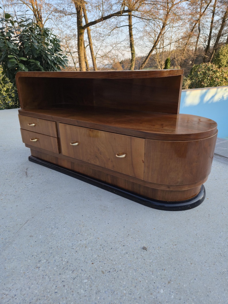 May include: A mid-century modern wooden cabinet with a curved design. The cabinet has a dark wood finish, three drawers with gold-colored handles, and a recessed top shelf. The base is black, and the piece is set on a concrete surface.