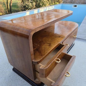 May include: A mid-century modern wooden cabinet with two drawers and a top shelf. The cabinet is made of a dark, glossy wood with a curved design. The drawers have gold-colored handles. The cabinet sits on a black base.