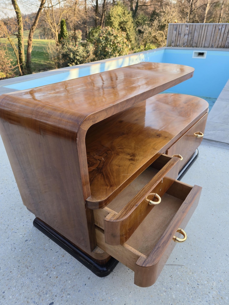 May include: A mid-century modern wooden cabinet with two drawers and a top shelf. The cabinet is made of a dark, glossy wood with a curved design. The drawers have gold-colored handles. The cabinet sits on a black base.