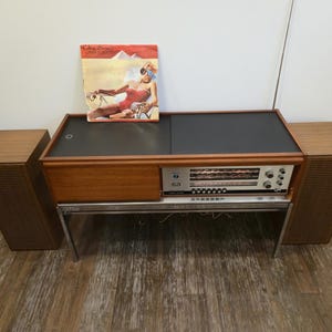 Puede incluir: Una consola estéreo vintage con mueble de madera, tapa negra y patas plateadas. La consola tiene una radio incorporada y altavoces. Una portada de álbum con una mujer en traje de baño rojo y gafas de sol descansa encima.