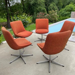 Peut inclure: Quatre chaises pivotantes avec un rev&ecirc;tement en tissu orange et des bases chrom&eacute;es. Les chaises sont dispos&eacute;es en cercle sur une terrasse en b&eacute;ton.