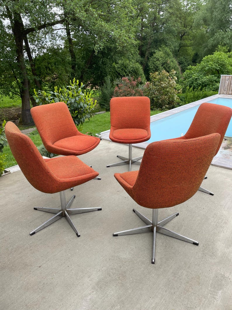 Peut inclure: Quatre chaises pivotantes avec un rev&ecirc;tement en tissu orange et des bases chrom&eacute;es. Les chaises sont dispos&eacute;es en cercle sur une terrasse en b&eacute;ton.