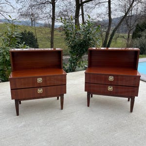 May include: Two matching wooden nightstands with two drawers each. The nightstands have a dark wood finish and gold-colored handles. The nightstands are sitting on a concrete floor.