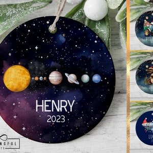May include: A round, blue and purple Christmas ornament with a watercolor galaxy design. The ornament features a yellow sun and planets in a line across the center. The text "HENRY 2023" is printed below the planets.