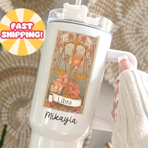May include: White tumbler featuring a Libra zodiac design with a balance scale, roses, and the name "Mikayla." The design is in soft pastel colors. The tumbler has a handle and a clear lid.