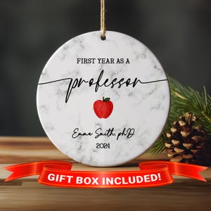 May include: White marble round ornament with the text "First Year as a Professor" in black cursive lettering. A red apple icon is centered below the text. The text "Emma Smith, PhD 2024" is below the apple icon.  "Gift Box Included!" is printed in red on a red ribbon.