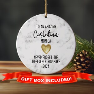 May include: White ceramic ornament with a marble pattern, featuring the text "To an amazing Custodian Monica Never forget the difference you make 2024". A gold heart with a gold outline is above the text "Monica". The ornament has a red ribbon and the text "Gift Box Included!" is at the bottom.