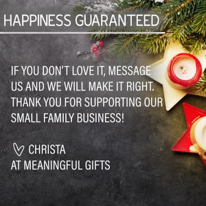 May include: A black background with a handwritten message that reads "Happiness Guaranteed. If you don't love it, message us and we will make it right. Thank you for supporting our small family business! &hearts; Christa at Meaningful Gifts". The message is surrounded by a pine branch with red berries and two gold star-shaped candle holders with red accents.