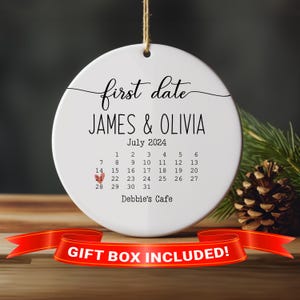 May include: White ceramic ornament with a calendar design featuring the text "first date" and the names "JAMES & OLIVIA". The calendar shows the month of July 2024 with a heart marking the 21st. The text "Debbie's Cafe" is printed below the calendar.