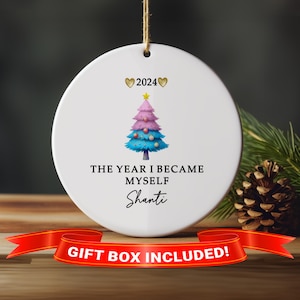 May include: White ceramic Christmas ornament with a colorful tree design. The tree is pink, blue, and green, with a gold star on top. The text reads "2024", "The Year I Became Myself", and "Shanti". A red ribbon with "Gift Box Included!" is at the bottom.