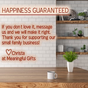 May include: A white ceramic mug with a black rim sits on a wooden surface. The background features a white brick wall with wooden shelves and greenery. The text "HAPPINESS GUARANTEED" is displayed in orange at the top of the image. The text "If you don't love it, message us and we will make it right. Thank you for supporting our small family business! Christa at Meaningful Gifts" is also displayed in orange.
