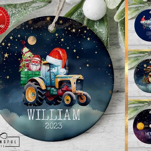 May include: A blue and gold Christmas ornament featuring a tractor pulling a Christmas tree with Santa Claus driving. The ornament has a white background with gold stars and the name "WILLIAM" and the year "2023" printed on it.