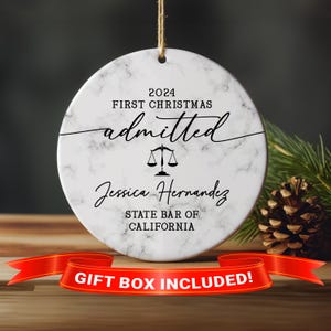 May include: A white marble round Christmas ornament with black text that reads "2024 First Christmas admitted" with a scale of justice icon and the name "Jessica Hernandez" and the text "State Bar of California".