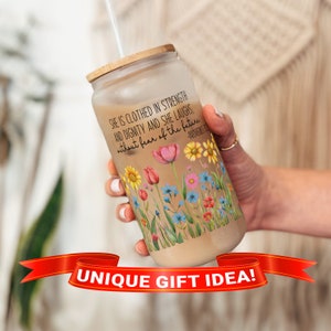 May include: A glass tumbler with a wooden lid and a straw. The tumbler has a floral design and a quote that reads "She is clothed in strength and dignity and she laughs, without fear of the future - Proverbs 31:25".